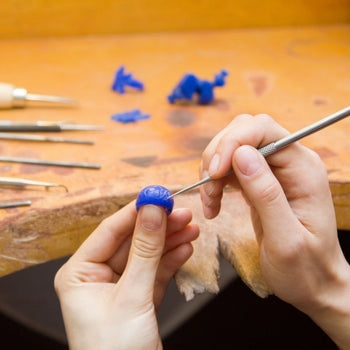 Hands carving intricate design on blue wax for jewelry making.